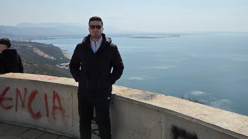 Hilltop scenic overlook with a curved concrete parapet and tiled pavement, looking out over a wide bay and distant coastline with hazy mountains under a clear sky.