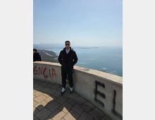 Hilltop scenic overlook with a curved concrete parapet and tiled pavement, looking out over a wide bay and distant coastline with hazy mountains under a clear sky.
