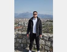 View from a stone lookout wall on a hillside overlooking a dense city with mid-rise buildings, with a rugged mountain range in the distance under a clear sky. Rocky terrain and sparse vegetation surround the viewpoint.