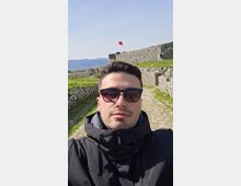 Stone fortress ruins with low stone walls and a cobblestone path, set on a grassy hillside with mountains in the distance. A red flag flies above the main stone rampart under a clear blue sky.