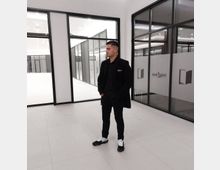 Bright, modern office or showroom corridor with white tile floors and black-framed glass walls and doors. Frosted glass panels display a logo and text, and large windows open to an interior courtyard with additional glass partitions.