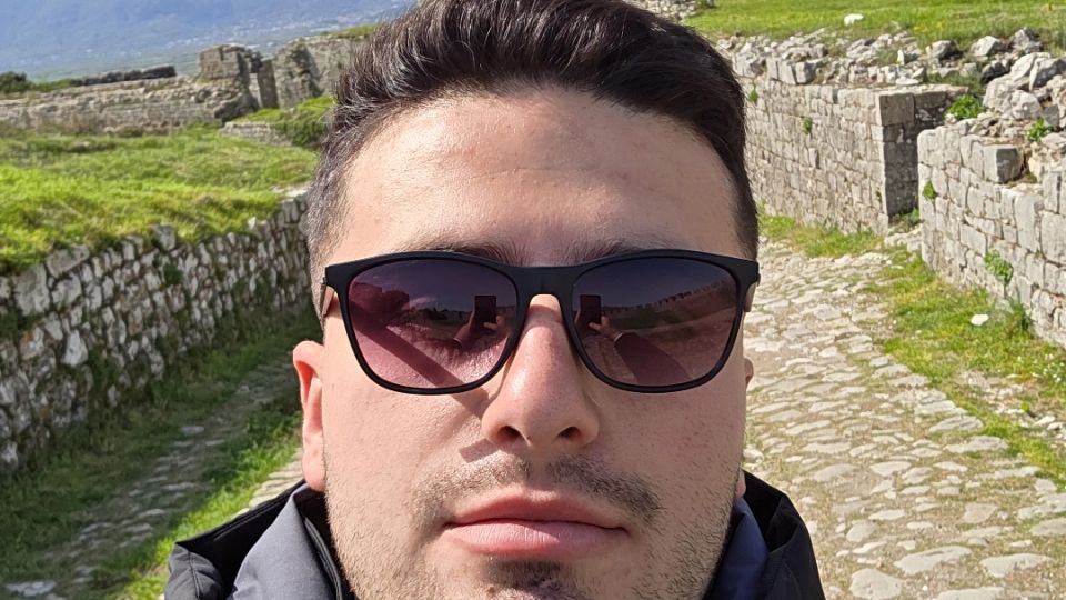 Stone fortress ruins with low stone walls and a cobblestone path, set on a grassy hillside with mountains in the distance. A red flag flies above the main stone rampart under a clear blue sky.