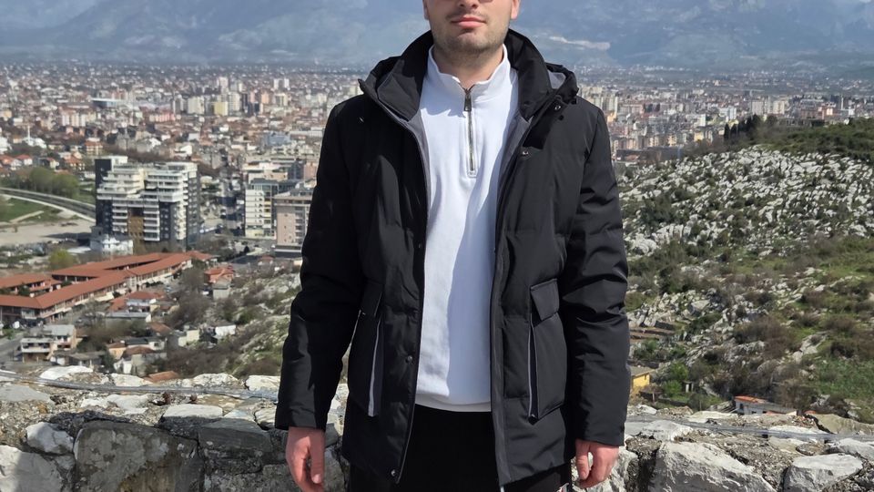 View from a stone lookout wall on a hillside overlooking a dense city with mid-rise buildings, with a rugged mountain range in the distance under a clear sky. Rocky terrain and sparse vegetation surround the viewpoint.